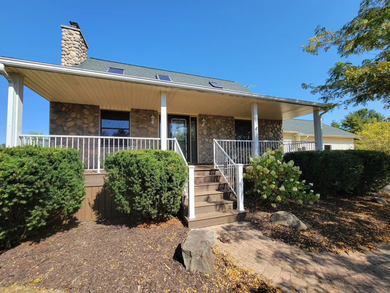 A covered front porch with brown decking, white railing, stone exterior walls, and stairs led to the house's entrance, surrounded by green shrubs.