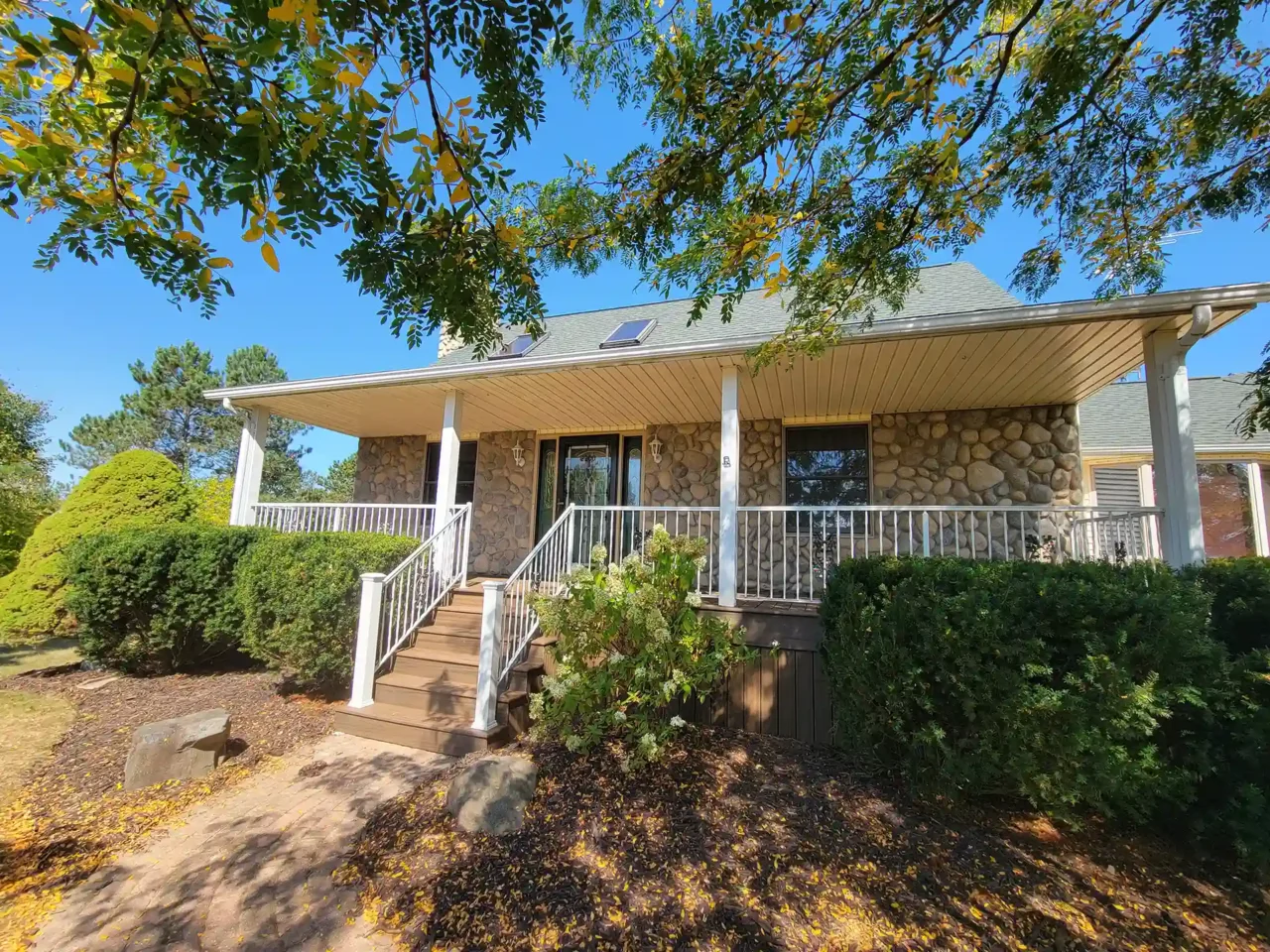 A covered front porch with brown decking, white railing, stone exterior walls, and landscaped bushes under a blue sky.
