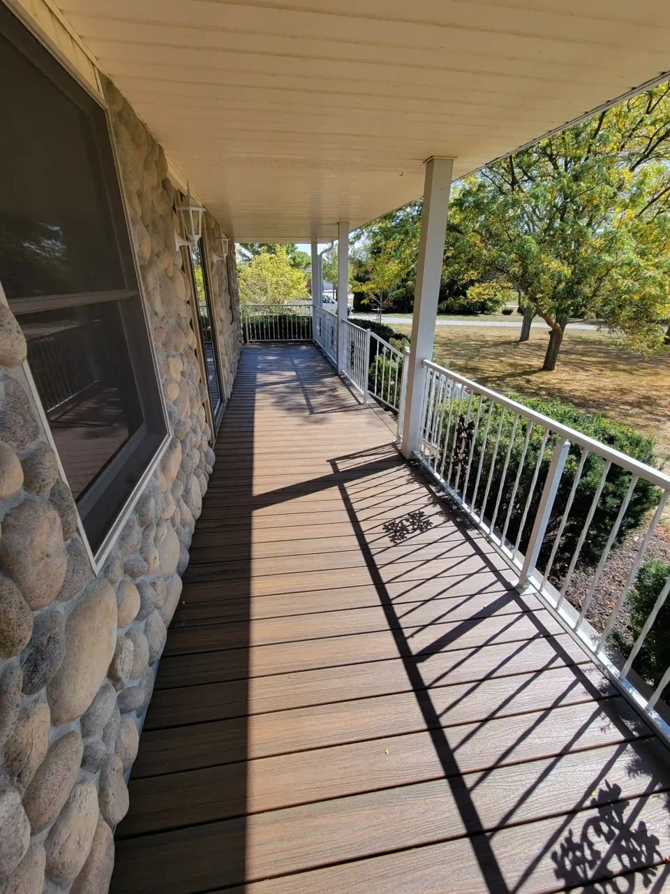 A covered porch with brown decking and white railing next to a stone exterior wall, overlooking a yard with trees.
