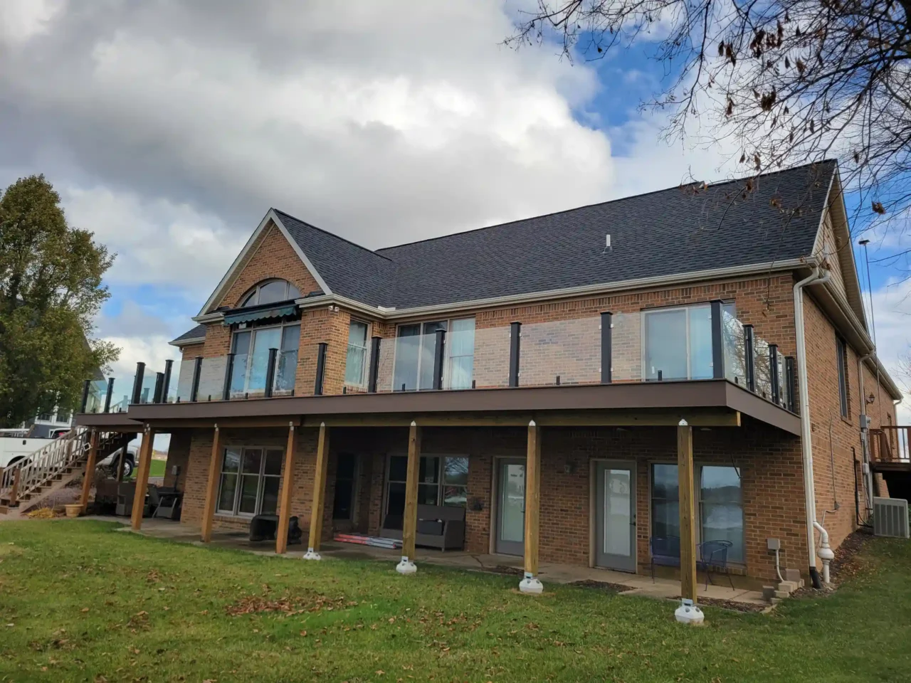 A large elevated deck with glass railing on a brick house, supported by wooden posts, overlooking a green lawn.