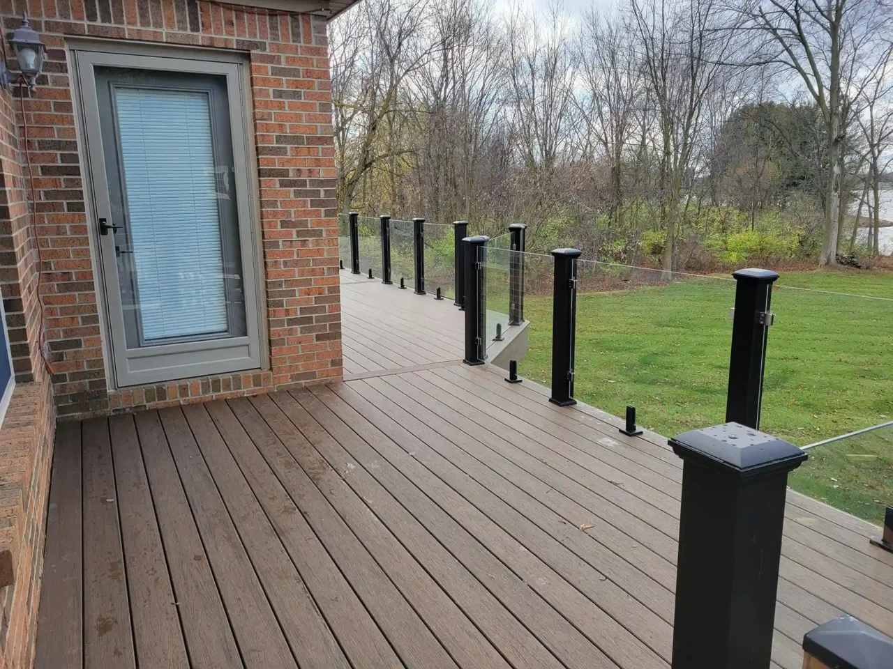 A brown composite deck with black glass railing posts attached to a brick house, overlooking a green backyard with trees.