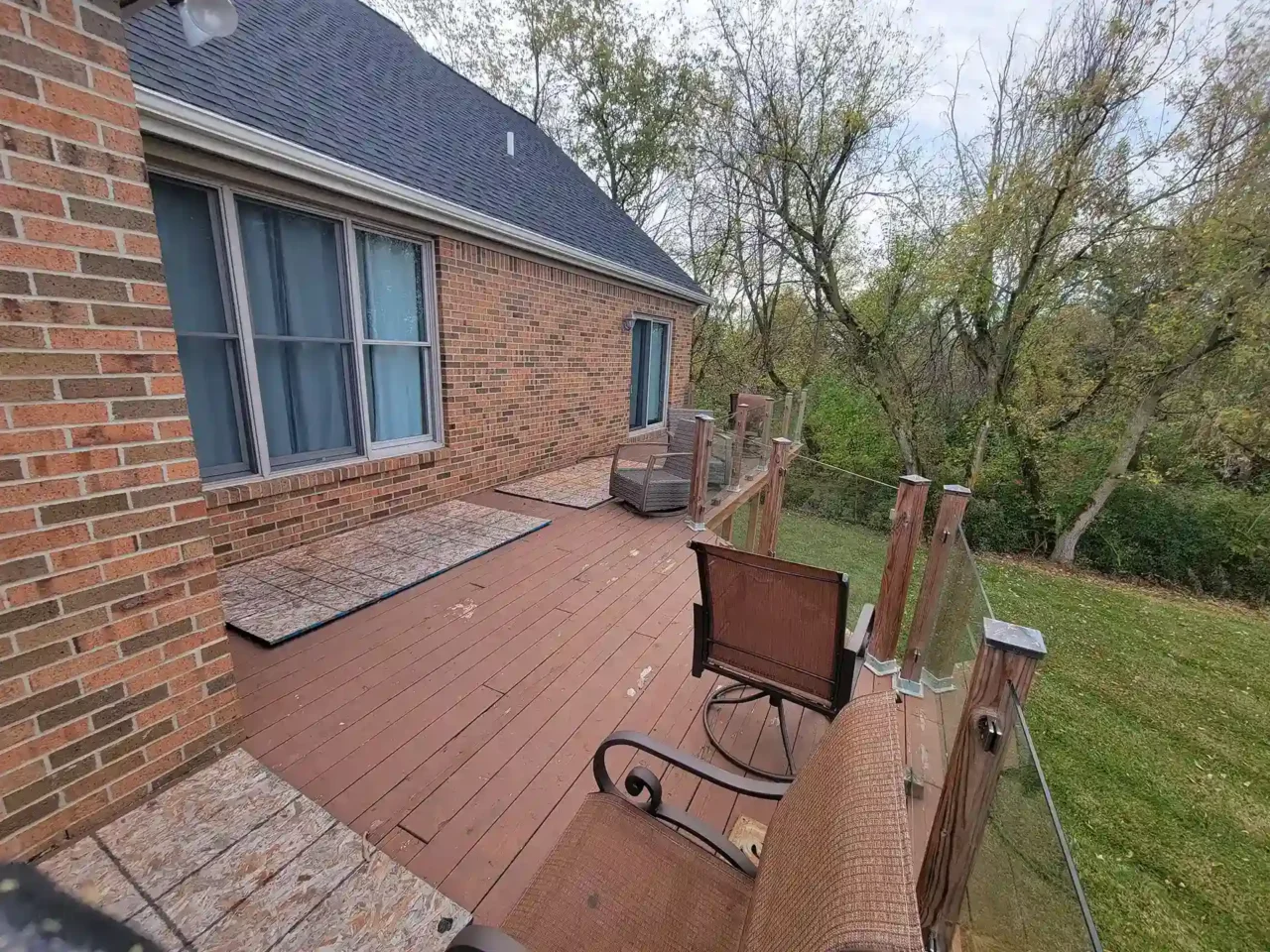 A smaller wooden deck with glass railing posts attached to a brick house, featuring outdoor seating and overlooking trees and grass.