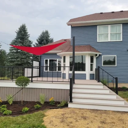 A deck with black metal railing and red shade sail is attached to a blue two-story house, surrounded by a garden with small plants and mulch.