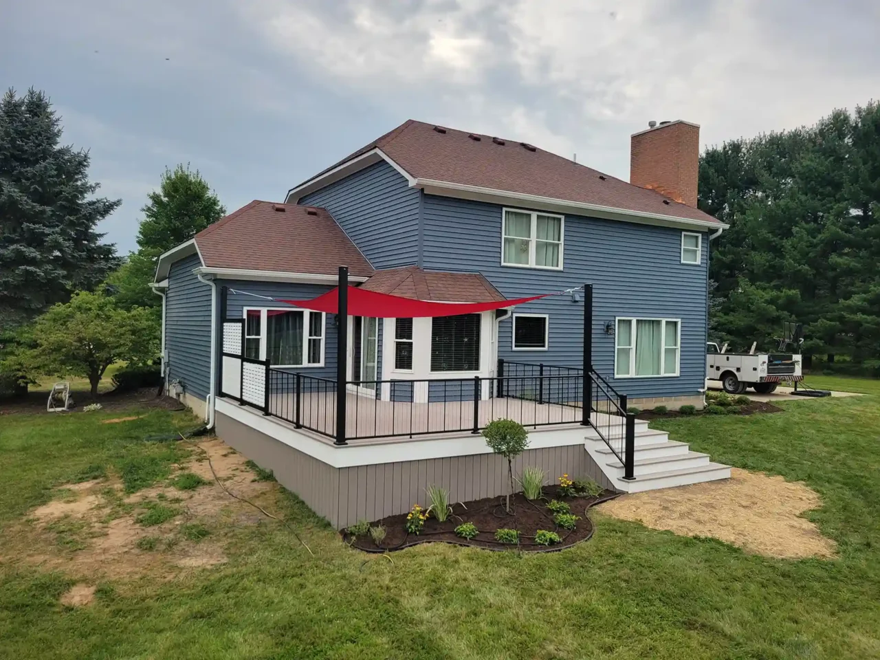 A newly built deck with black metal railing and red shade sail is attached to a blue house, surrounded by a lawn and small landscaped flower beds.