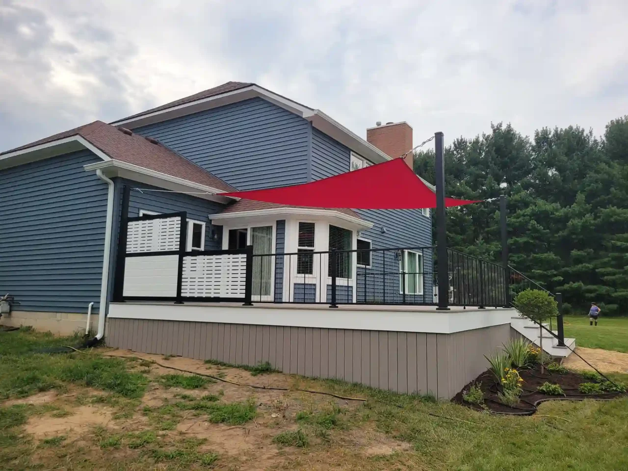 Elevated deck with black metal railing and red shade sail viewed from the side, attached to a blue house with multiple windows and white trim.