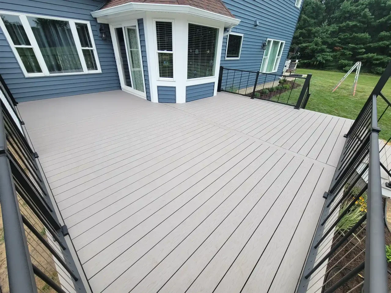 Spacious beige composite deck with black metal railing attached to a blue house featuring a white bay window, overlooking a green lawn.