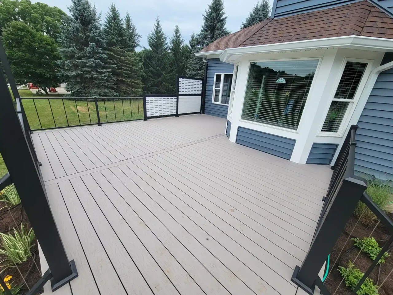 Beige composite deck with black metal railing and privacy screen, attached to a blue house with a white bay window, overlooking a landscaped yard and pine trees.