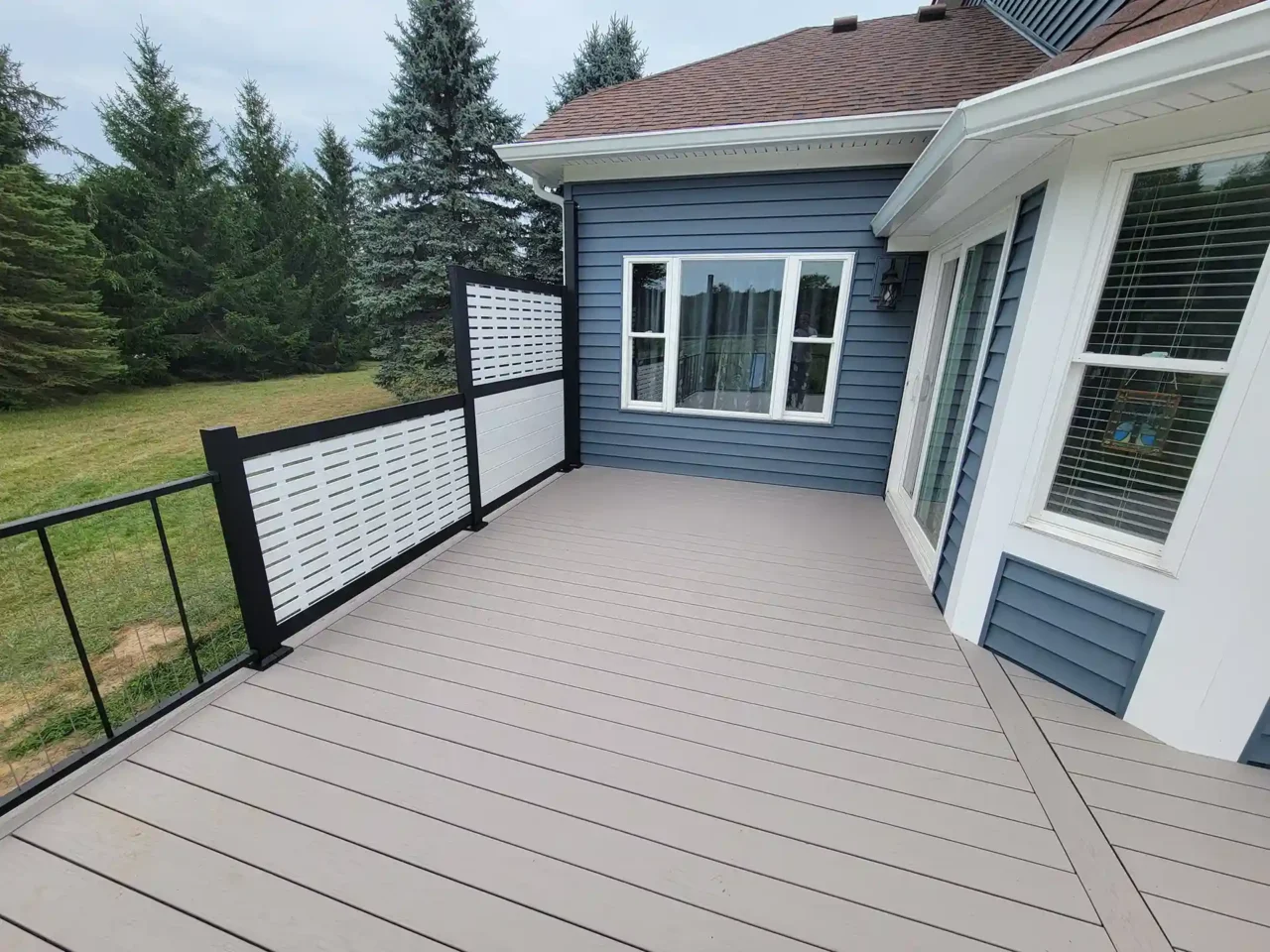 Beige composite deck with black metal railing and white privacy screen attached to a blue house with multiple windows, overlooking a grassy yard bordered by trees.