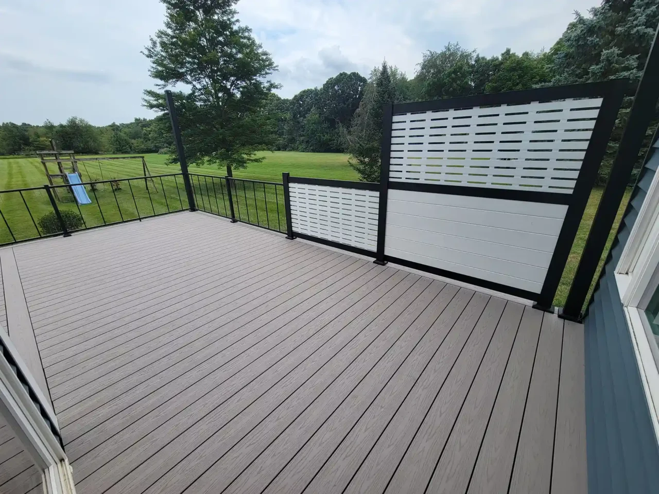 Beige composite deck with black metal railing and white privacy panels, overlooking a large grassy backyard with trees and a children’s swing set.