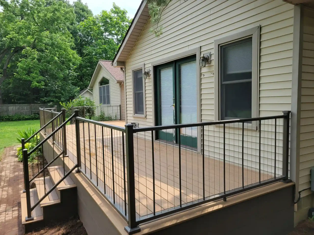 Side view of an elevated composite deck with TimberTech Antique Leather decking and black cable railing, attached to a beige house with green French doors, surrounded by lush greenery.
