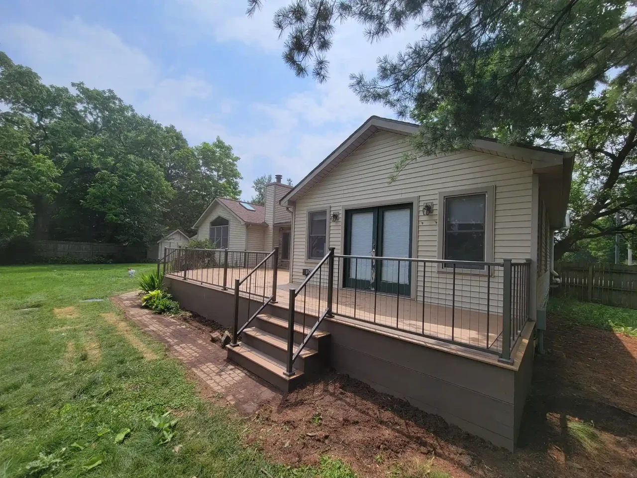 Angled view of a composite deck with TimberTech Antique Leather decking and black cable railing, attached to a beige house with green French doors.