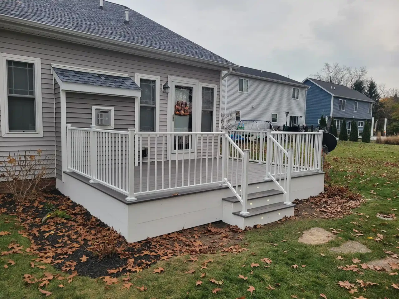 A gray deck with white railing and stairs leading to a backyard next to a light gray house on an overcast day.