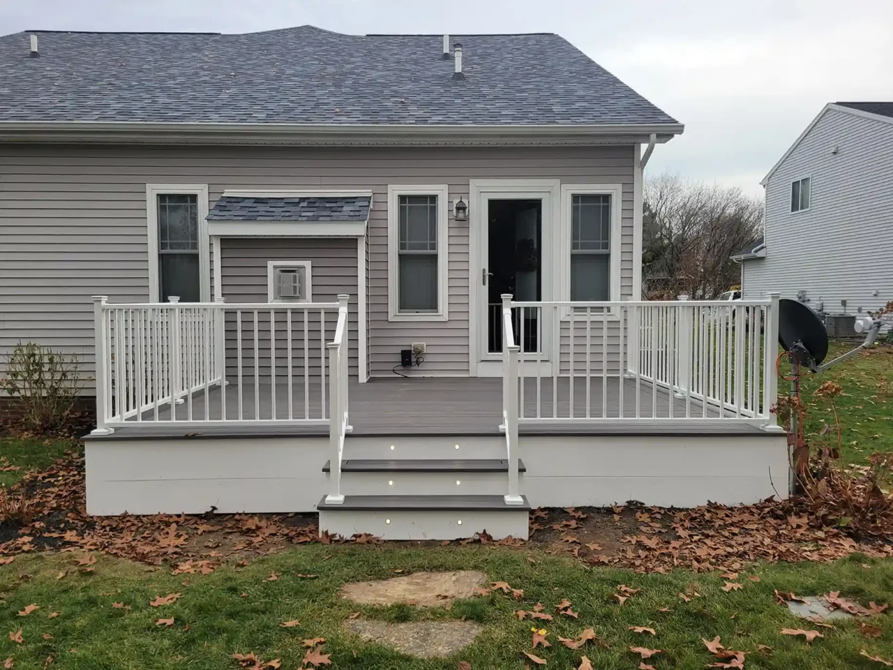 A gray PVC deck with white railing and illuminated steps is attached to the back of a light gray house.