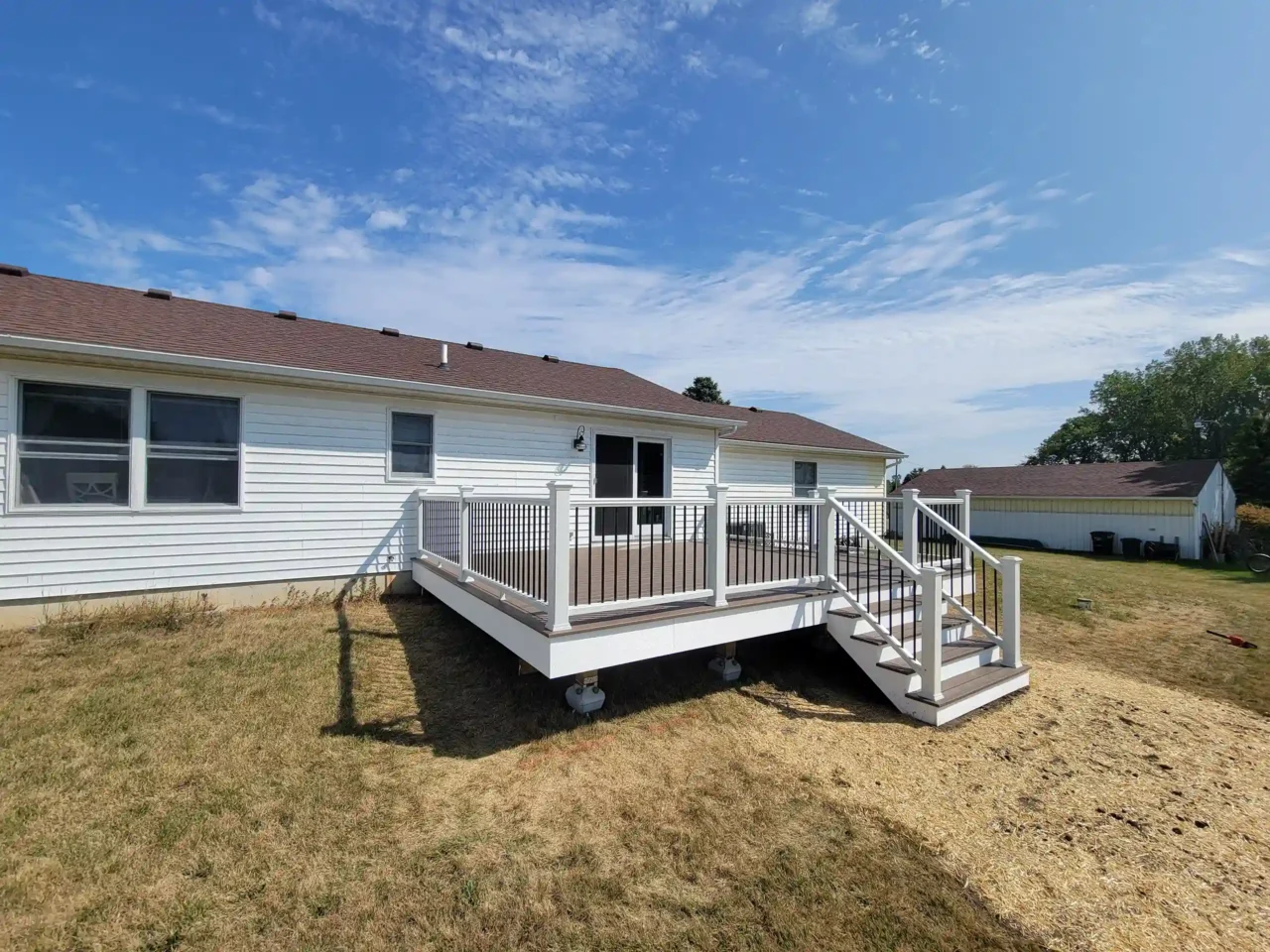 Newly built elevated deck with white vinyl railing and black balusters attached to a white single-story house with a brown roof.