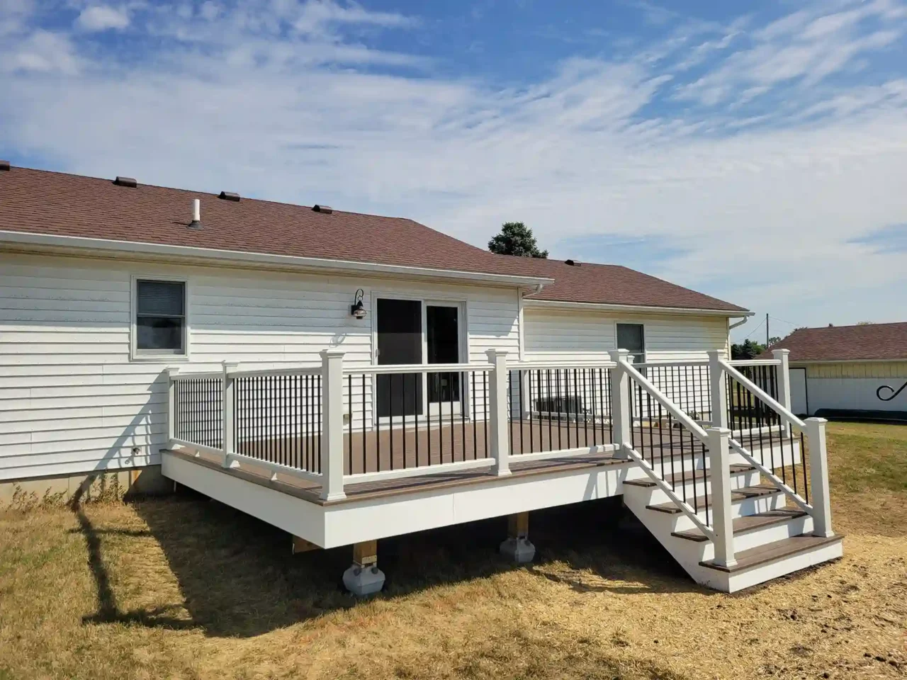 Elevated deck with white vinyl railing and black balusters attached to a white house with a brown roof, surrounded by dry grass under a partly cloudy sky.