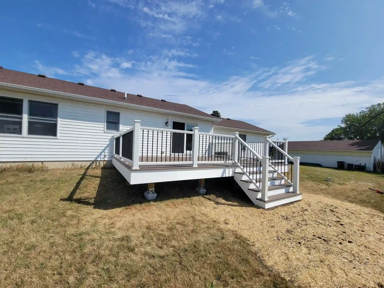 Elevated deck with white vinyl railing and black balusters attached to a white house with a brown roof, surrounded.