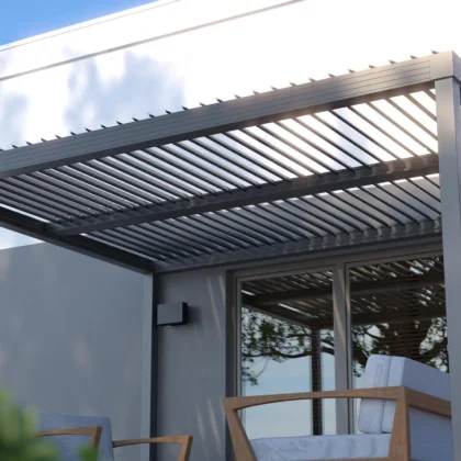 A modern pergola attached to a white building with large glass sliding doors. The pergola is dark gray with horizontal slats on top. Two outdoor chairs with white cushions are visible beneath it.