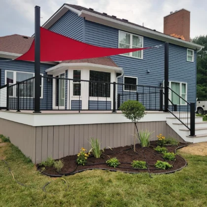 Back of a two-story house with blue siding and a brown roof. A large, multi-level deck extends from the house, featuring black railings and white posts. Part of the deck is shaded by a red triangular sun sail.