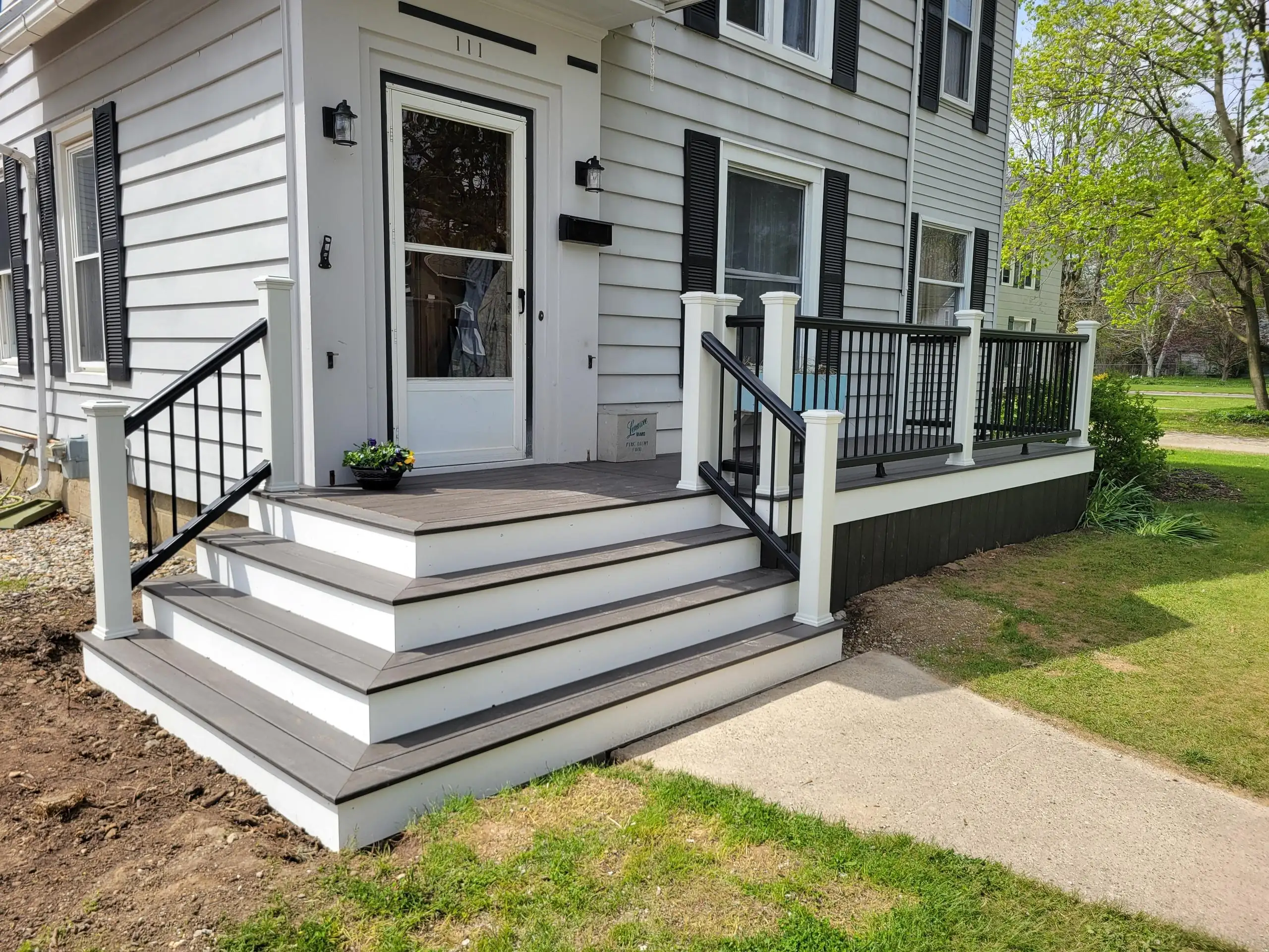 PVC vs Composite Decking A close-up view of a new front porch and stairs attached to a light gray or white house with black shutters. The stairs and porch decking are made of dark gray composite material, framed by white risers, stringers, and posts. The railings are a combination of white posts and black metal handrails and balusters.